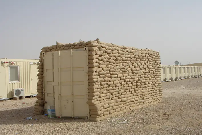 Pramo Prefabrik askeri konteyner, güvenlik amaçlı kum torbalarıyla çevrilmiş şekilde sahada konumlandırılmış dayanıklı modüler yapı olarak görülüyor.